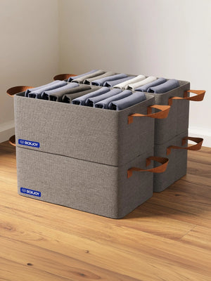 Four grey BOXJOY stackable fabric storage boxes filled with folded clothes on a wooden floor.